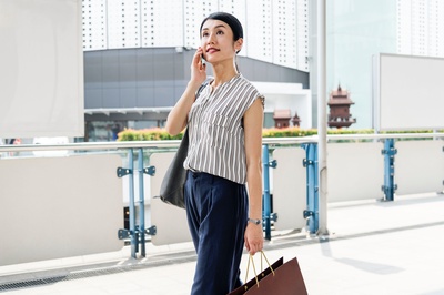 购物到下车的图片-穿棕色和白色细条纹无袖上衣的女人拿着纸手提袋，用智能手机在白色建筑物附近。